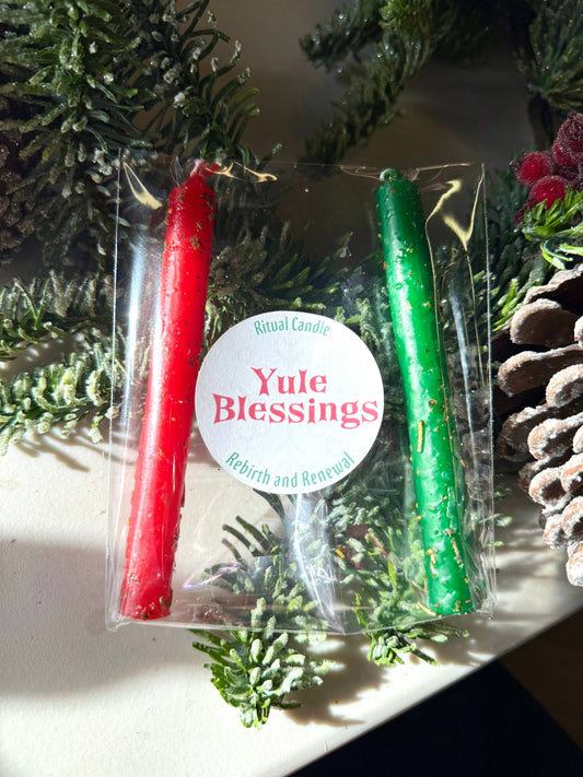 One red and one green taper candle with a Yule Blessings Ritual Candle label among winter greenery