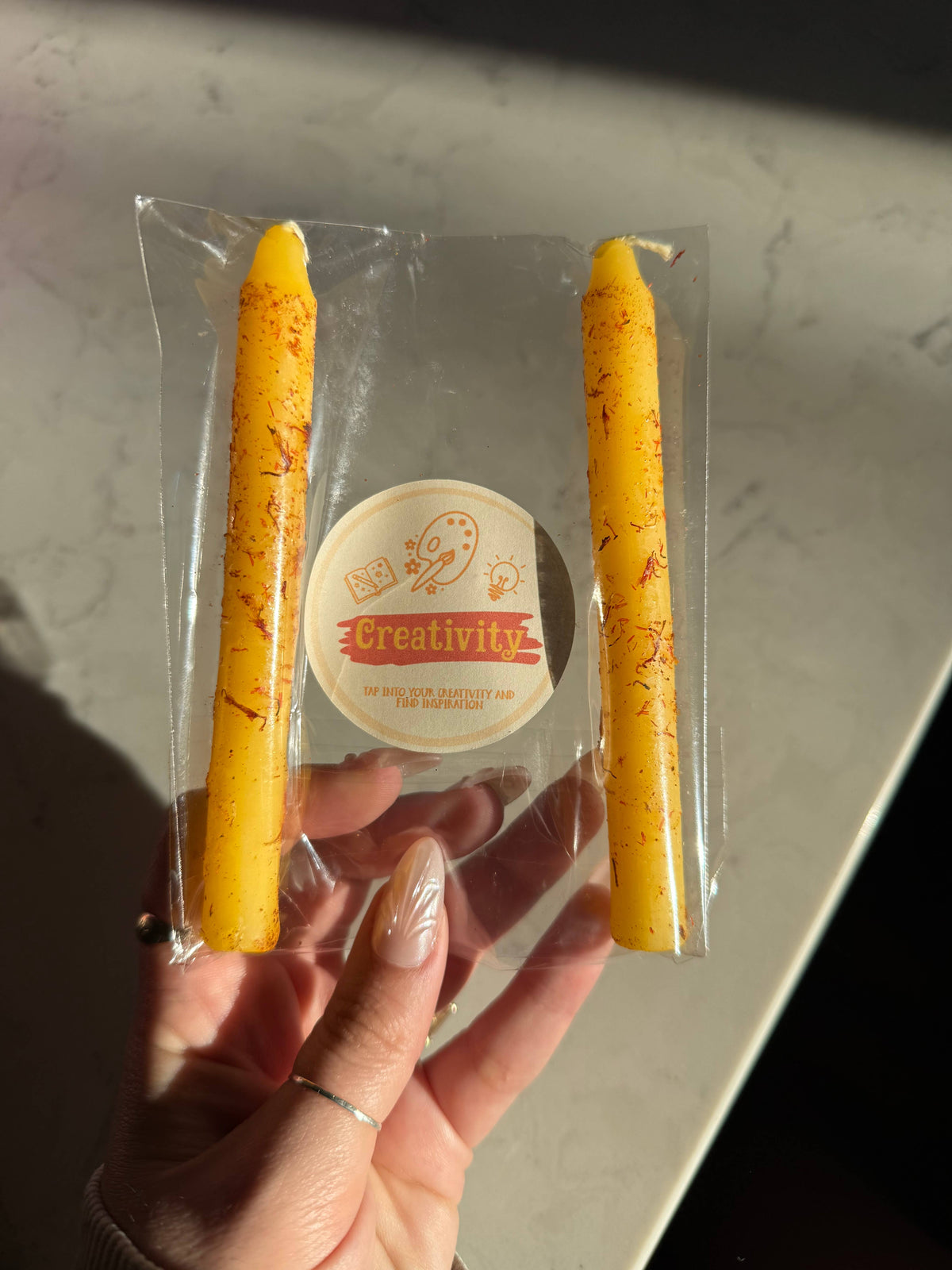 Two golden yellow taper candles in a clear sleeve with a Creativity Ritual Candle label