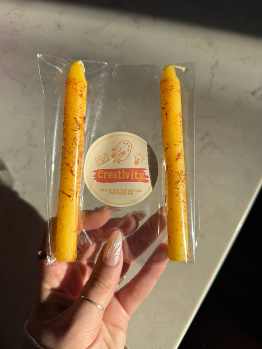 Two golden yellow taper candles in a clear sleeve with a Creativity Ritual Candle label
