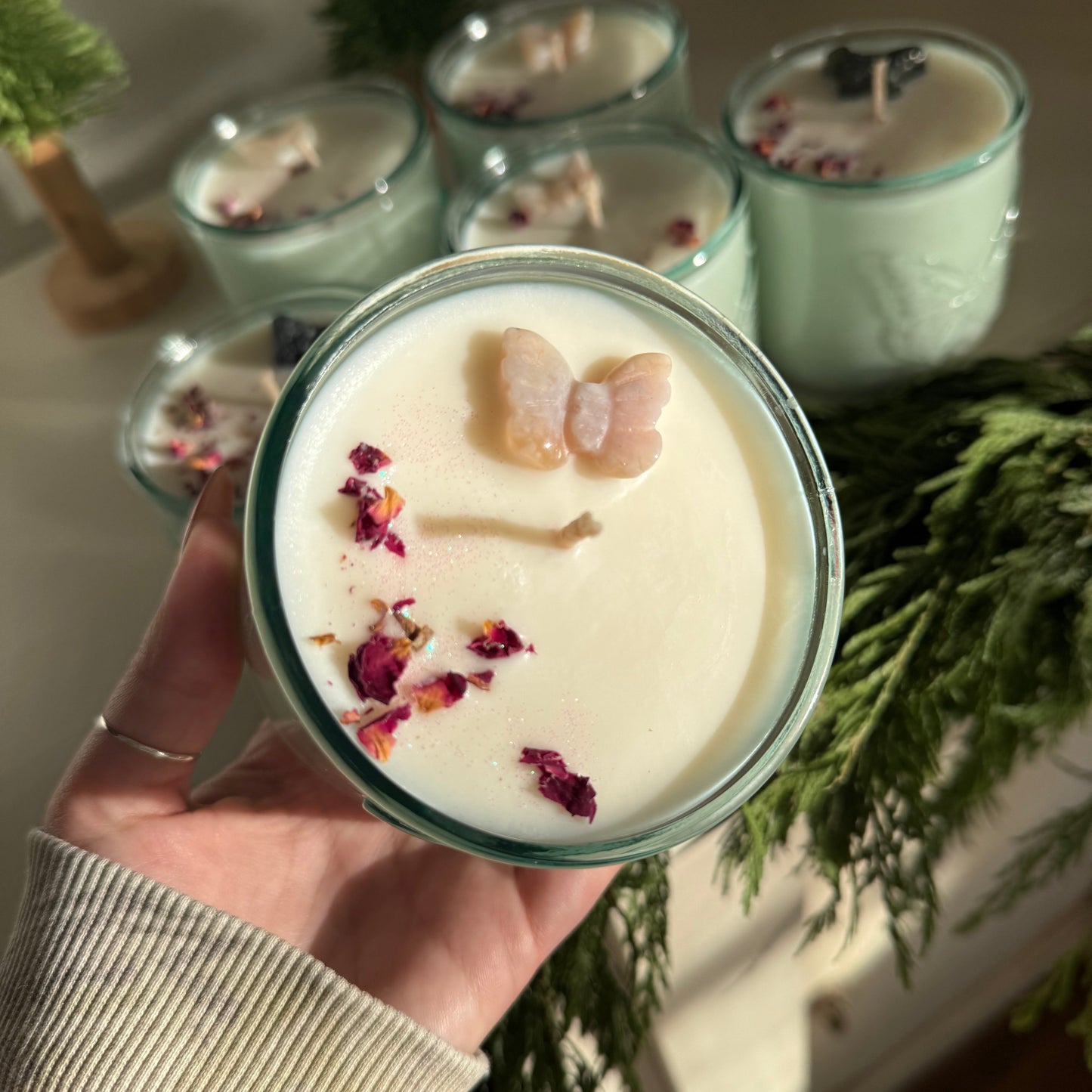 Hand holding a candle with decorative elements, surrounded by more candles and greenery.