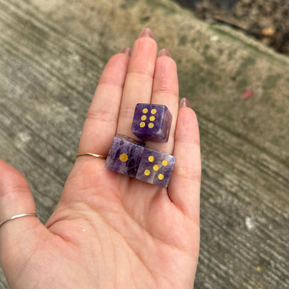 Hand holding two purple dice with gold pips on a concrete surface