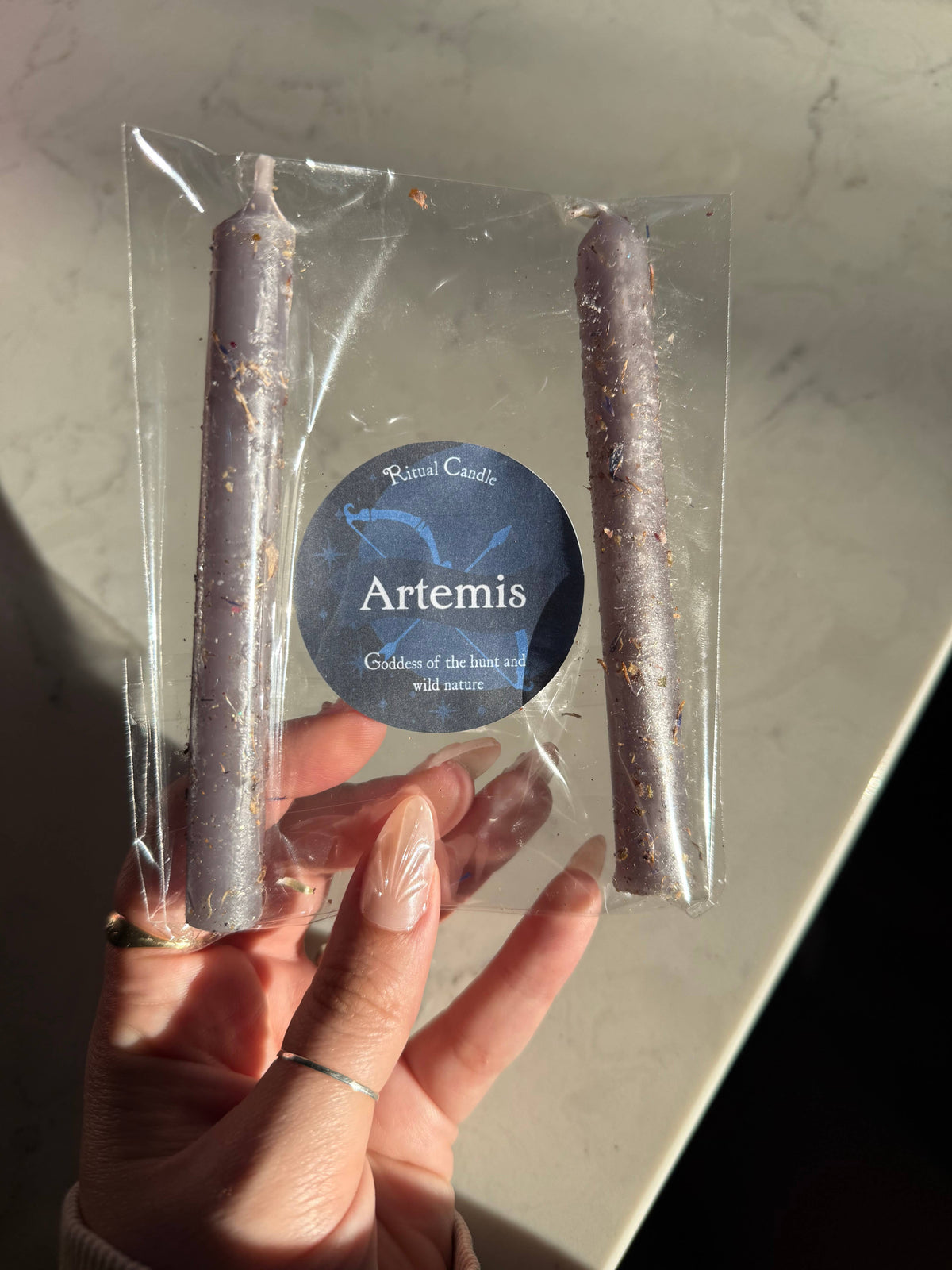 Two grey taper candles in a clear sleeve with a blue Artemis Ritual Candle label