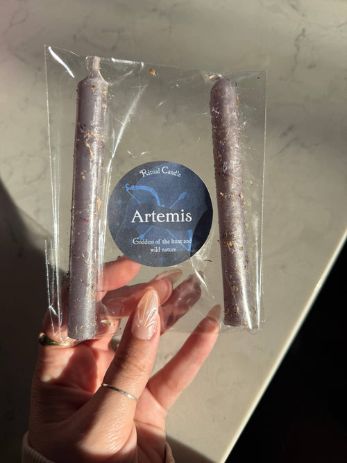 Two grey taper candles in a clear sleeve with a blue Artemis Ritual Candle label