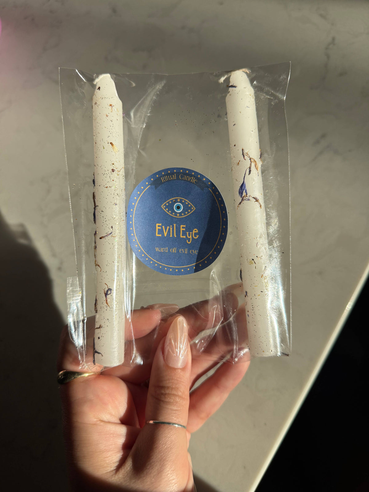Two white taper candles in a clear sleeve with a blue Evil Eye Ritual Candle label